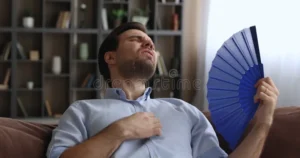 tired overheated young man cooling himself wave hand handheld fan suffers summer unbearable heat home sweaty guy sit 221707220